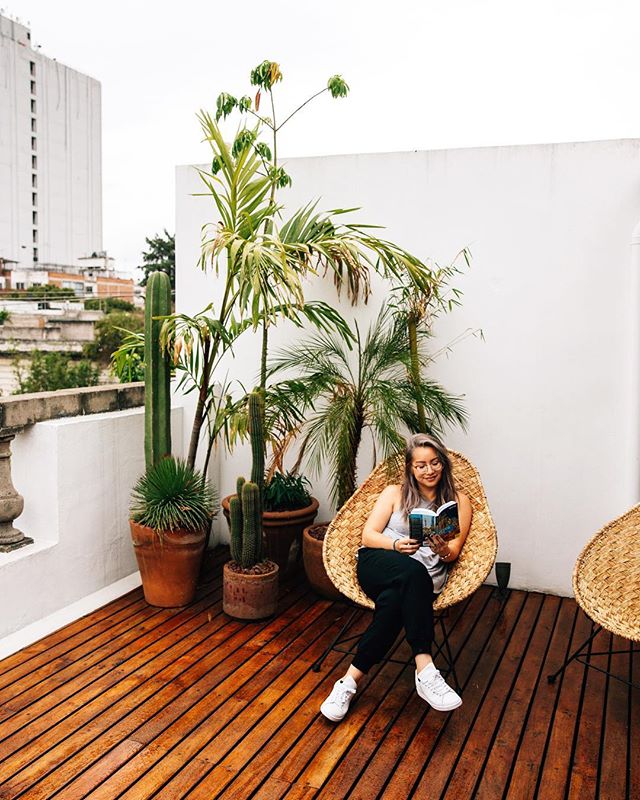 This is a photo of a girl sitting on a deck in a straw chair reading a book with large green plants behind her.