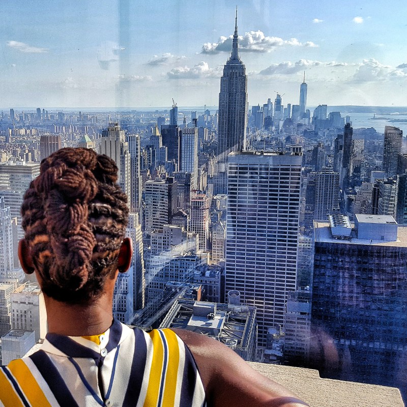 A photo of a girl looking out through glass at the Empire State Building and the Lower Manhattan Skyline.