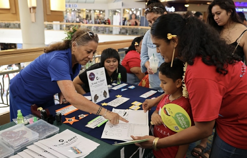 Tapati, pictured far right, is shown helping her son with a language exercise during a Mahina ʻŌlelo Hawaiʻi community event. Rise